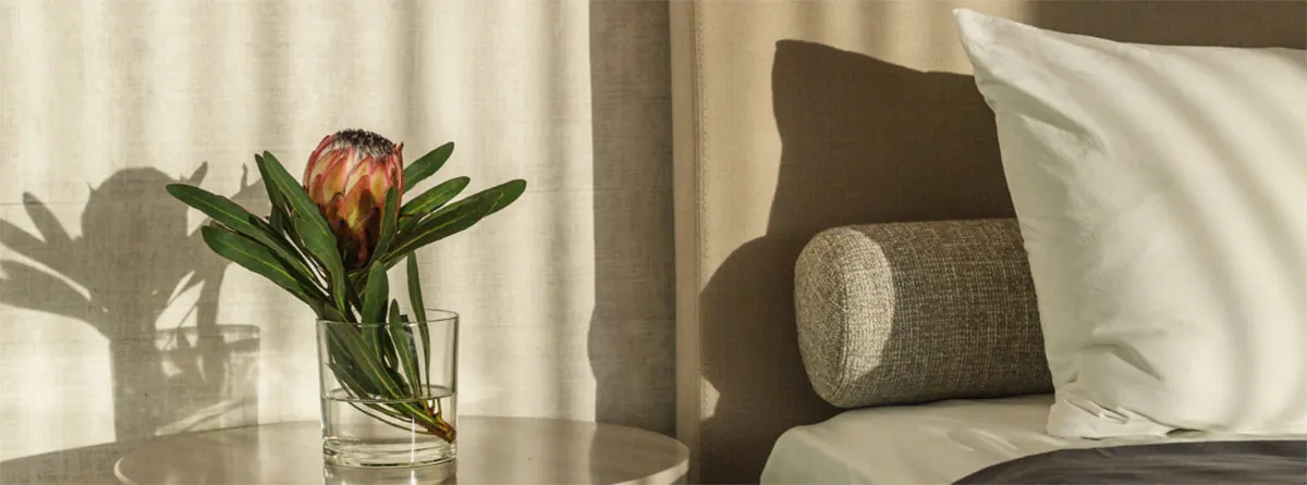 Tavolino con un fiore di protea in un bicchiere accanto al letto, con cuscino bianco e cuscino cilindrico in tessuto