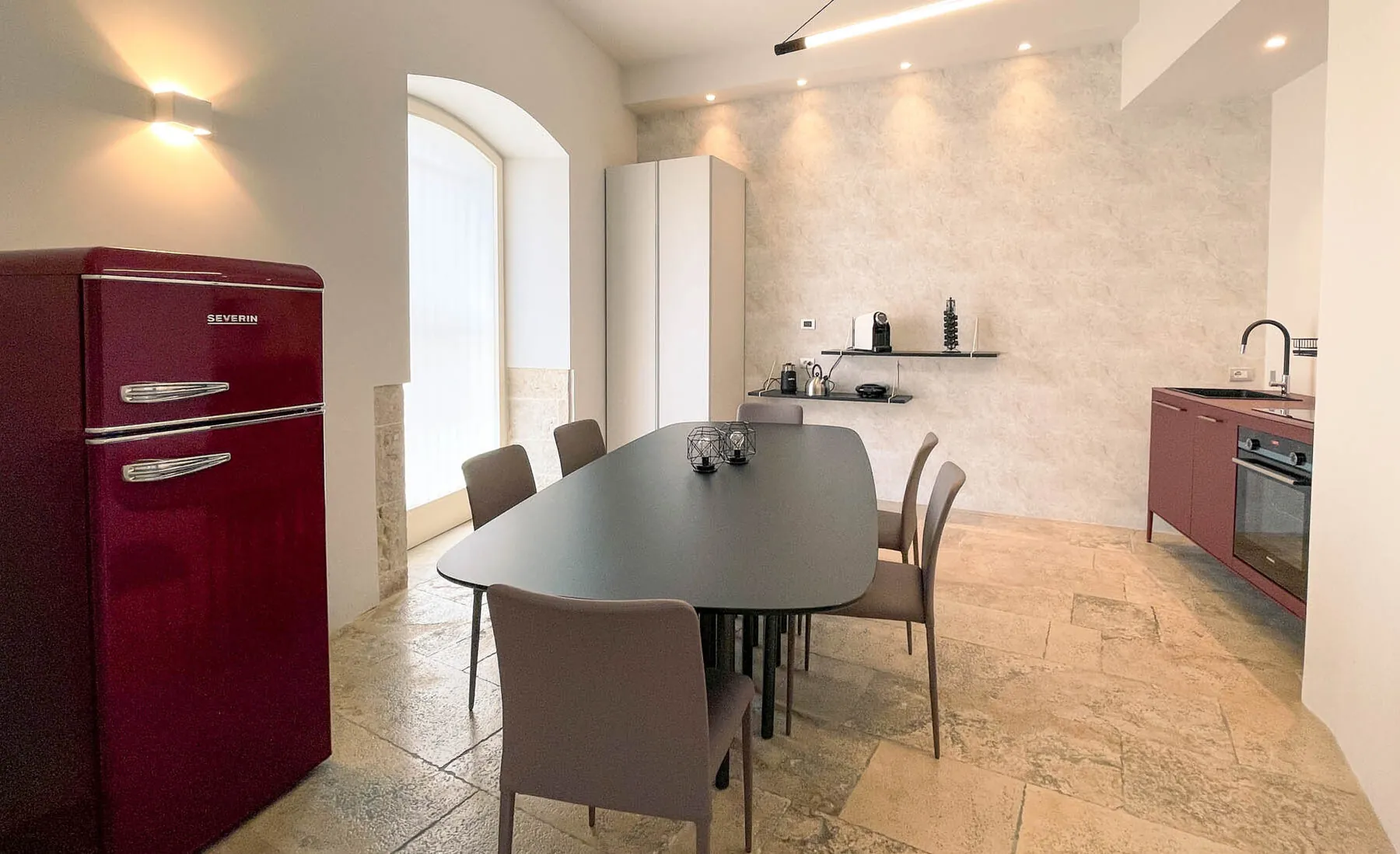 La cucina comune con tavolo lungo e sedie, frigorifero rosso in stile anni Cinquanta e finestra ad arco