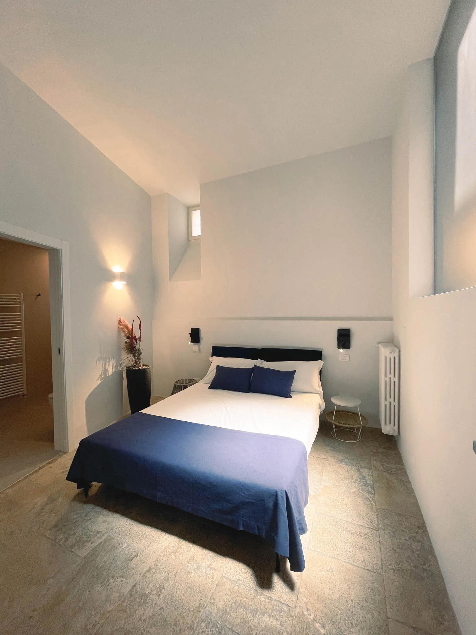 Il letto matrimoniale della camera Pètra con copriletto blu e applique sulla parete bianca
