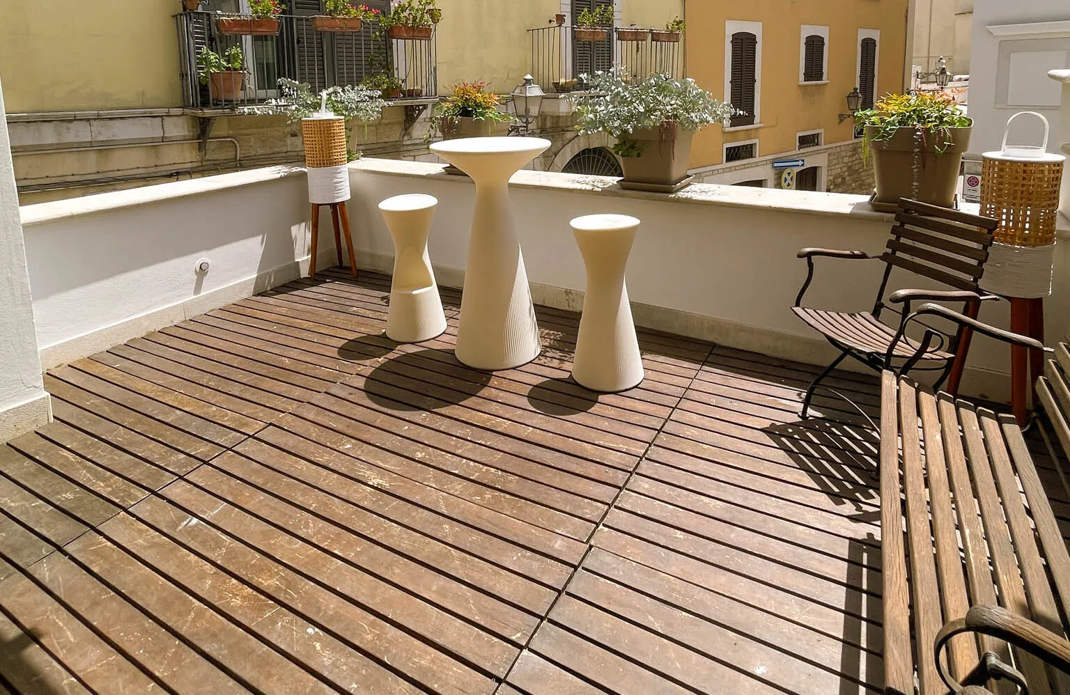 Terrazzino con tavolini alti bianchi e panca in ferro sulla pedana in legno, fra i balconi fioriti del vicolo