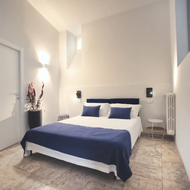 Camera Pètra: letto matrimoniale con copriletto blu, pareti bianche e pavimento in pietra chiara