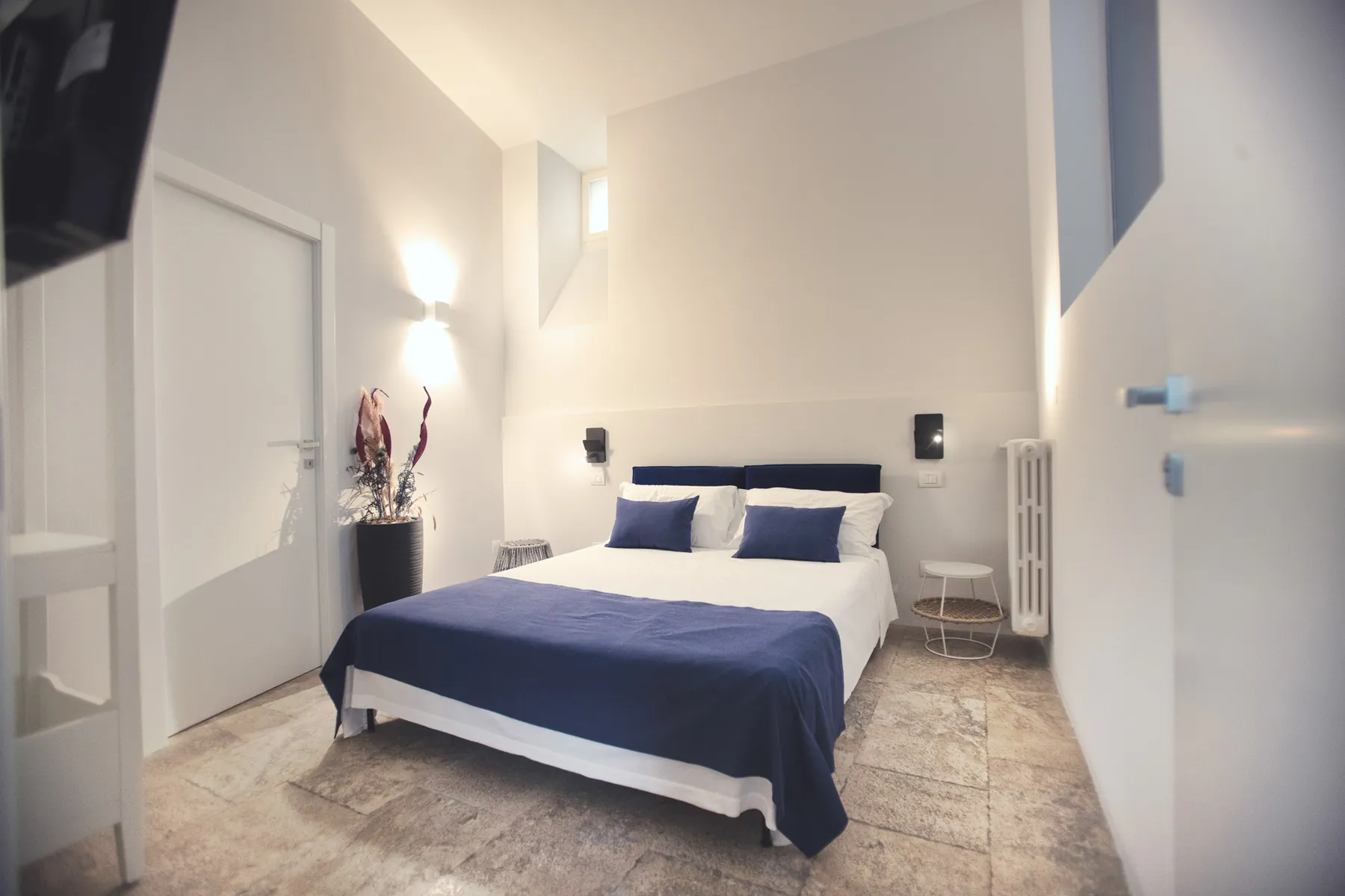 La camera Pètra: letto matrimoniale con testiera e copriletto blu, pareti bianche e pavimento in pietra chiara