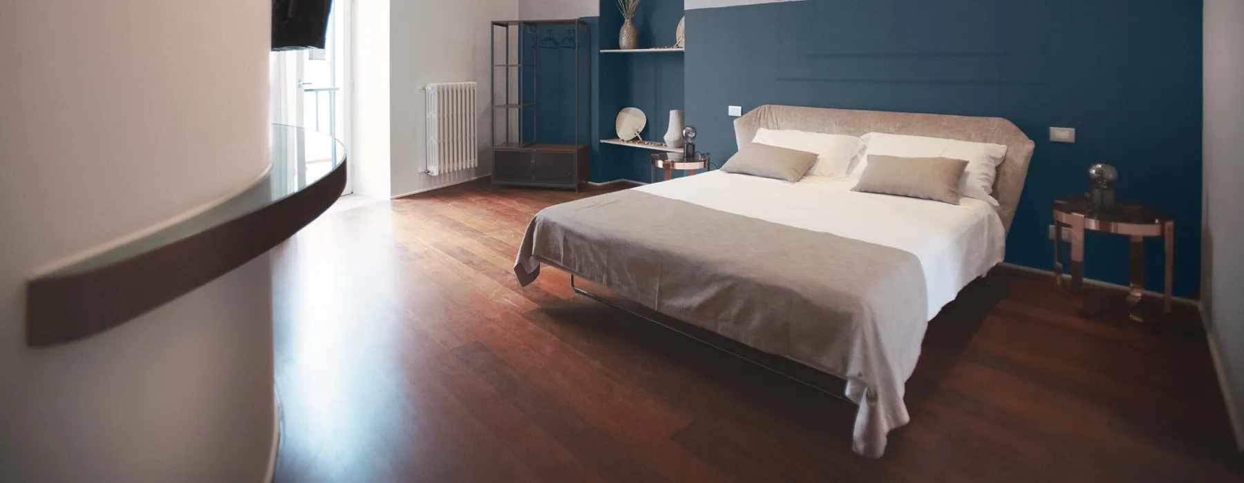 La camera Xylo: letto matrimoniale bianco davanti alla parete blu petrolio, pavimento in legno scuro